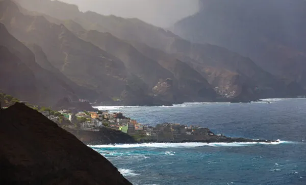Een spectaculair gezicht op een dorpje gelegen op een rotsachtige kaap aan de wilde oceaan onder een dramatische bergketen op Santo Antão, Kaapverdië.