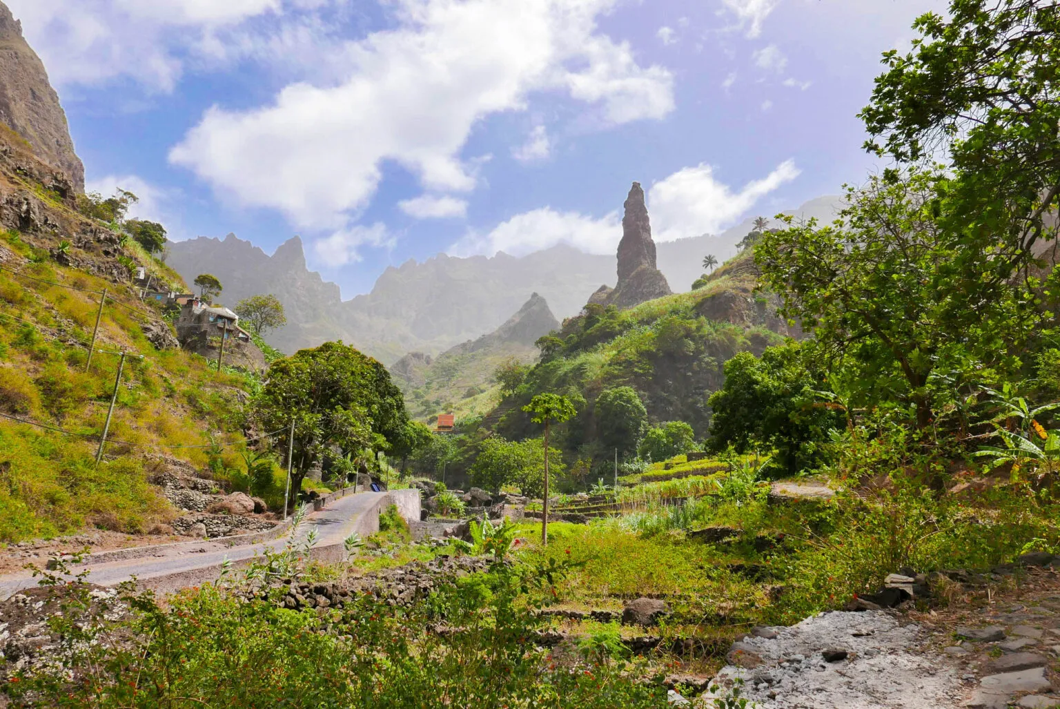 Een groene bergvallei in Xoxó op Santo Antão met een opvallende, steile rotsnaald en een slingerende weg onder een lichtbewolkte blauwe lucht.