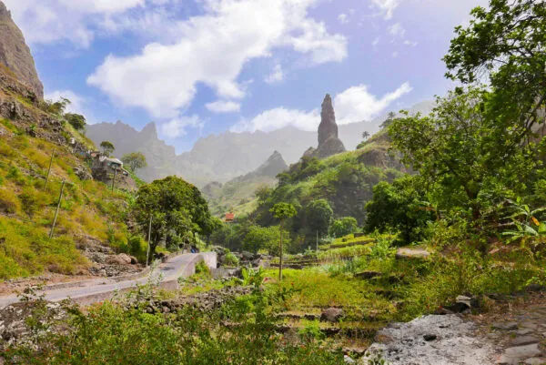 Een groene bergvallei in Xoxó op Santo Antão met een opvallende, steile rotsnaald en een slingerende weg onder een lichtbewolkte blauwe lucht.