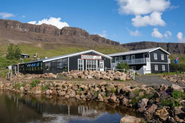Zicht op het zwarte houten gebouw van Hraunsnef accommodatie aan een klein waterbed met rotsachtige oever, met op de achtergrond een steile, groene bergwand onder een lichtbewolkte blauwe lucht in IJsland.