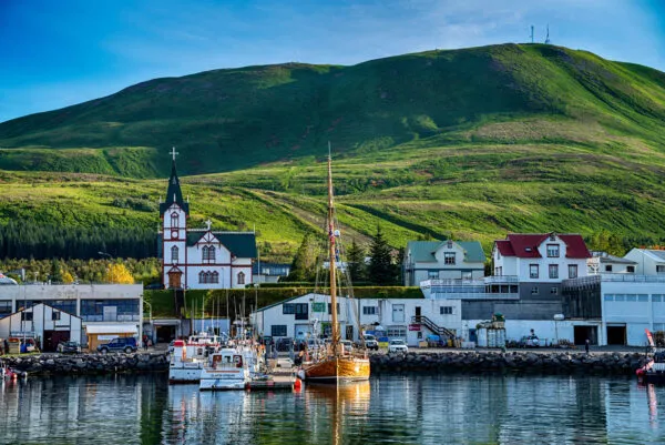 Zicht vanaf het water op de schilderachtige haven van Húsavík in IJsland, met traditionele houten (walvis)boten aan de kade, de karakteristieke houten kerk Húsavíkurkirkja en een grote groene berg op de achtergrond.