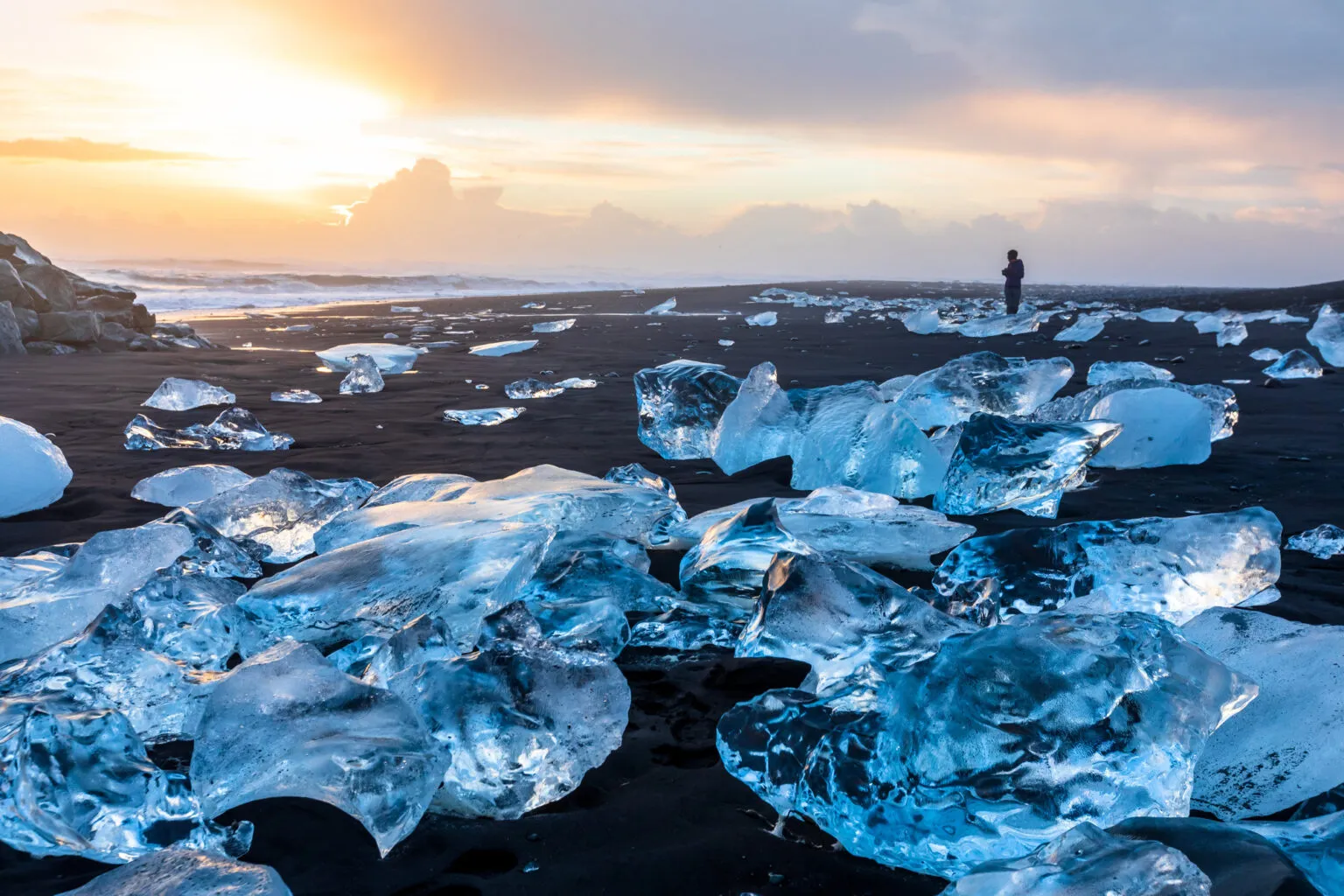 Transparante, glinsterende blokken gletsjerijs liggen verspreid op het zwarte vulkanische strand van Diamond Beach in IJsland, met de opkomende of ondergaande zon die een warme gloed over de oceaan en de horizon werpt.