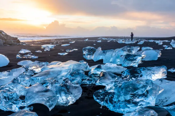 Transparante, glinsterende blokken gletsjerijs liggen verspreid op het zwarte vulkanische strand van Diamond Beach in IJsland, met de opkomende of ondergaande zon die een warme gloed over de oceaan en de horizon werpt.