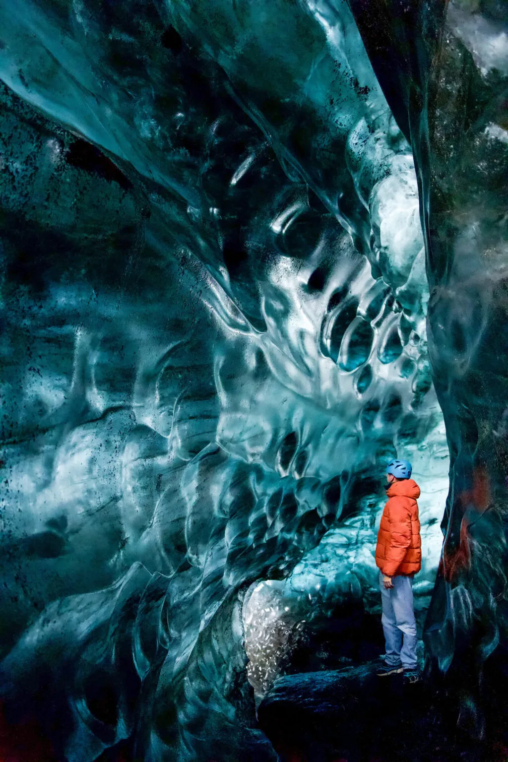 Een reiziger in een oranje jas wandelt door een indrukwekkende blauwe ijsgrot nabij de Jökulsárlón gletsjerlagune in IJsland.