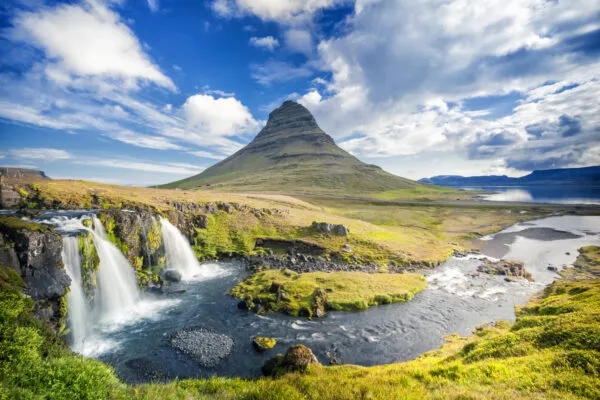 Zicht op de iconische, kegelvormige berg Kirkjufell op het schiereiland Snæfellsnes in IJsland, met de stromende Kirkjufellsfoss-waterval op de voorgrond onder een blauwe lucht met dynamische wolken.