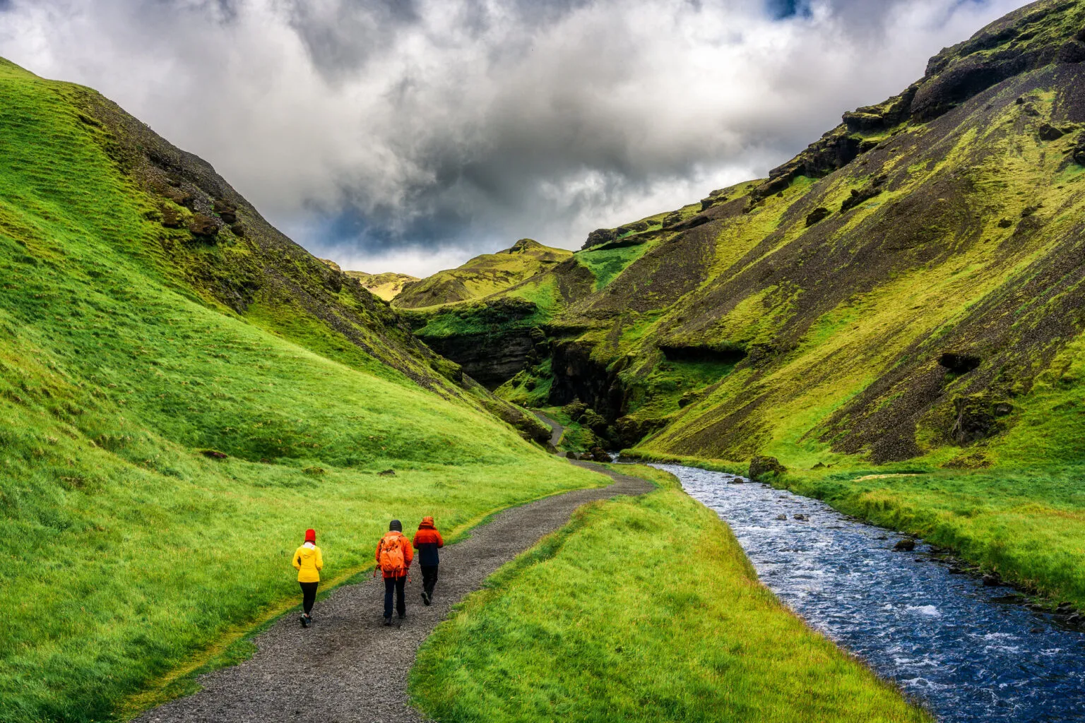 Een groep reizigers wandelt over een grindpad door een groene kloof langs een rivier richting de Kvernufoss waterval in IJsland.