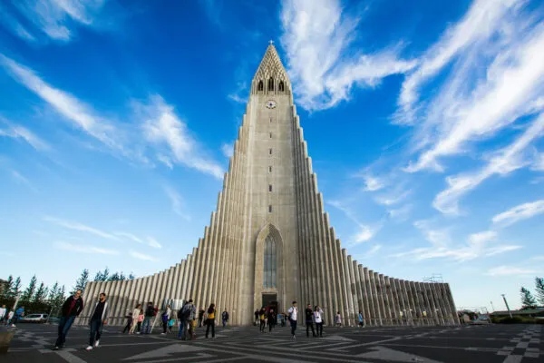 Kikvorsperspectief van de iconische betonnen voorgevel en toren van de Hallgrímskirkja-kerk in Reykjavik, IJsland, met bezoekers op het voorplein onder een heldere blauwe lucht met lichte bewolking.