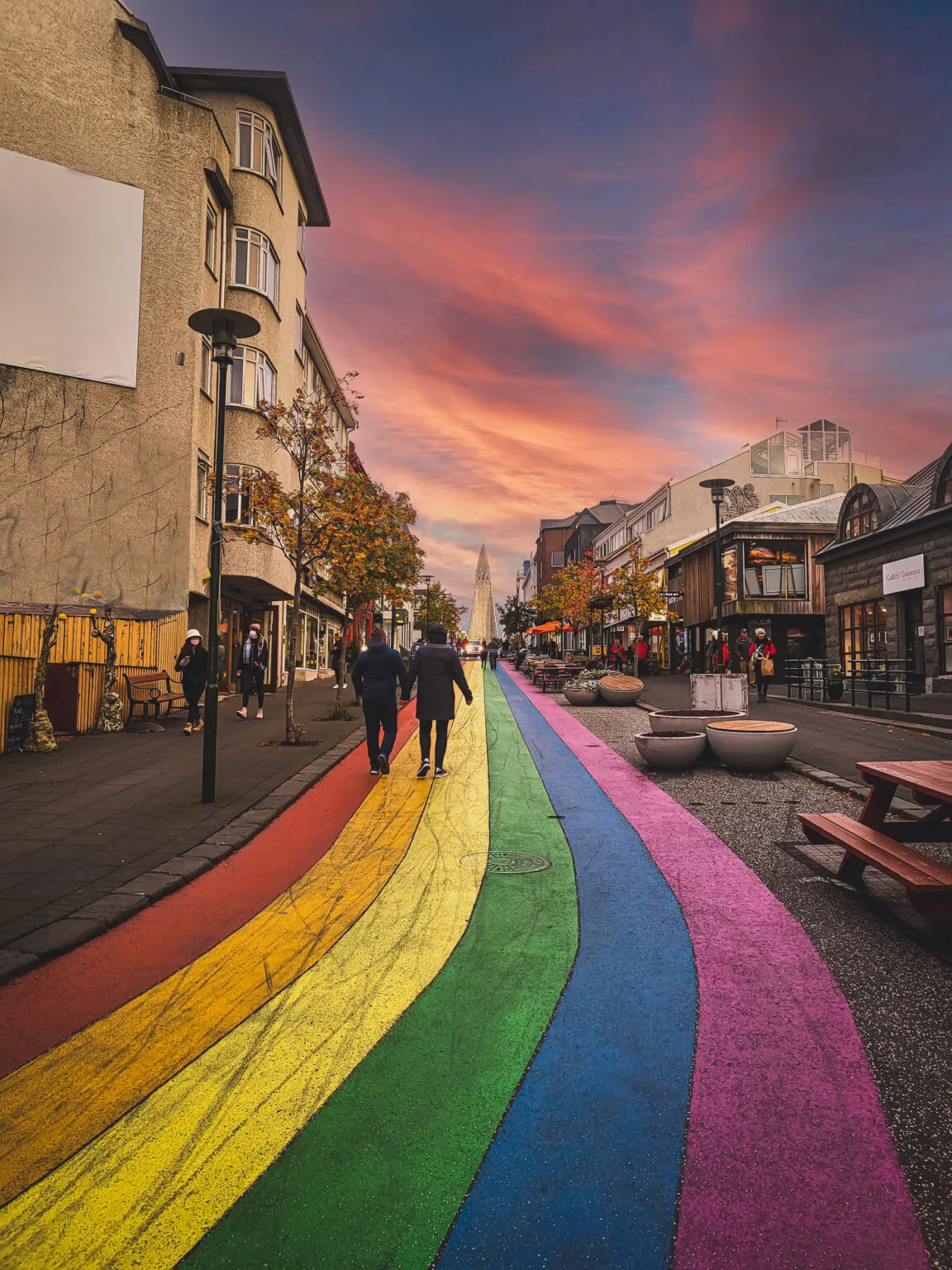 Perspectief over het kleurrijke regenboogpad in de straat Skólavörðustígur in Reykjavik, met wandelende mensen en een kleurrijke avondlucht op de achtergrond.