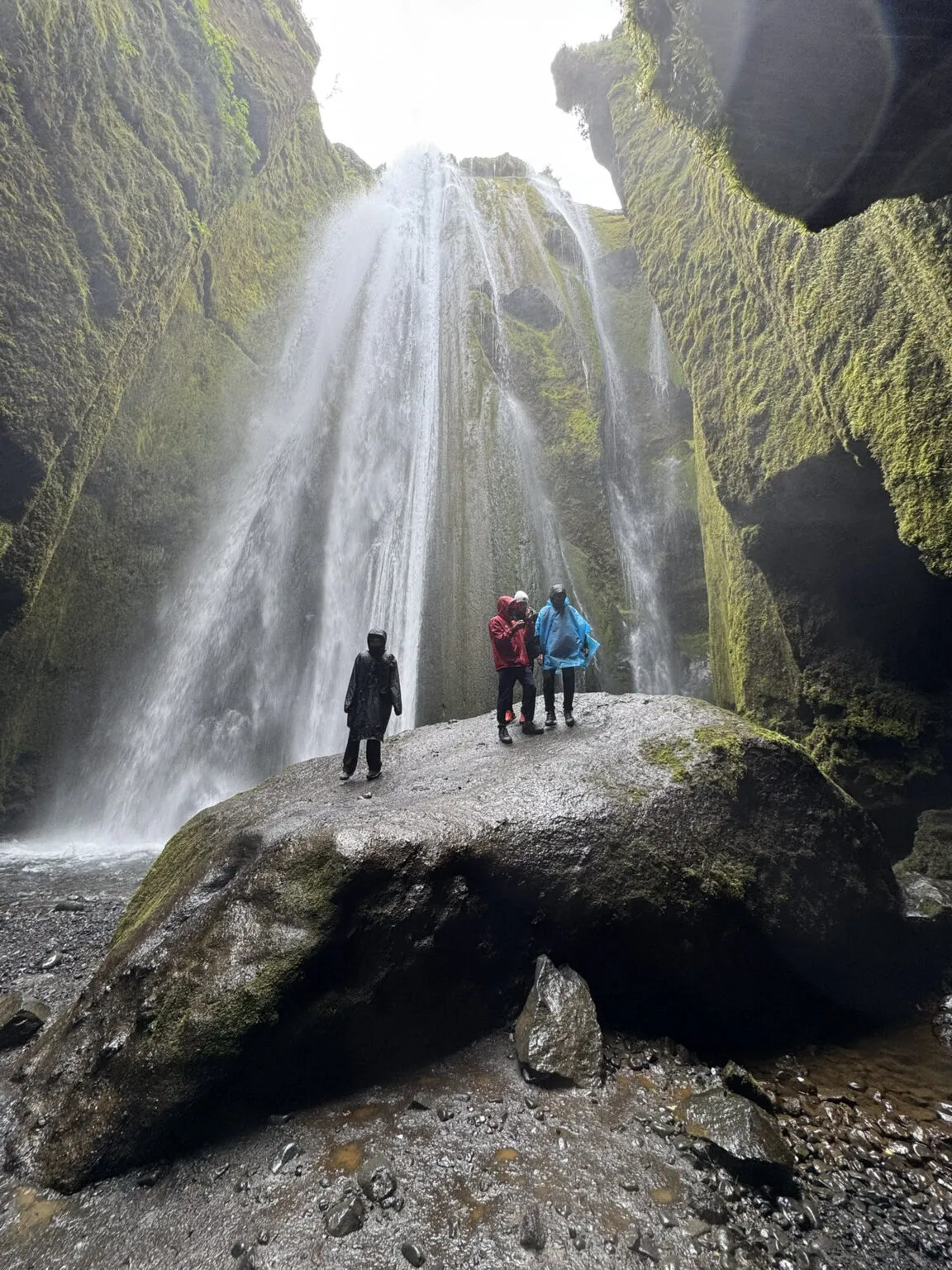 Wandelaars staan op een grote rots in een smalle, mosbegroeide rotskloof terwijl de verborgen Gljúfrabúi-waterval krachtig achter hen naar beneden stort in IJsland.