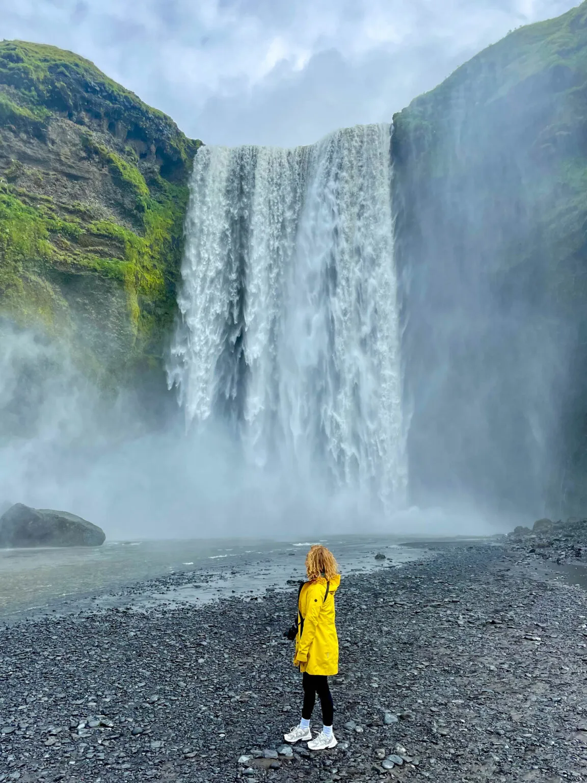 Een reiziger in een opvallende gele jas staat op het zwarte kiezelstrand en kijkt op naar de grote, krachtige Skógafoss-waterval die van een mossige klif naar beneden stort in Zuid-IJsland.