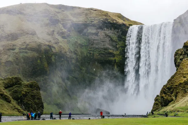 Zicht op de indrukwekkende Skógafoss-waterval in IJsland, waar een brede watermassa naar beneden stort vanaf een steile, groenbewassen klif met reizigers die aan de voet van de waterval staan.