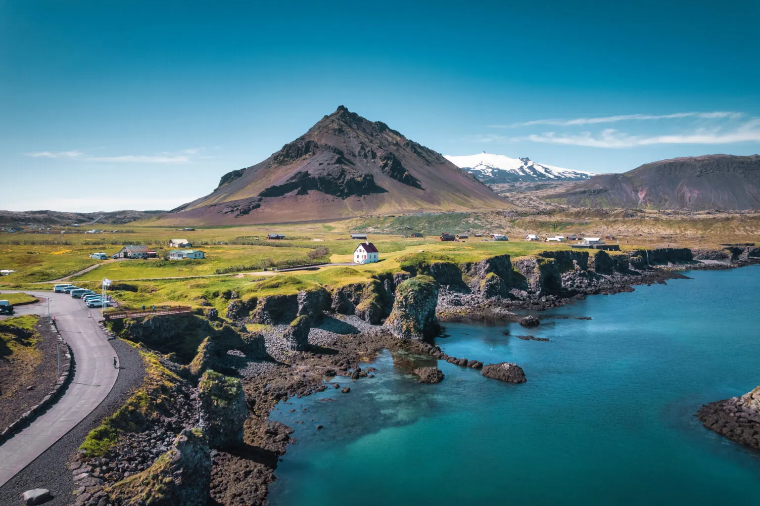 Weids panormagewelf over het kustdorp Arnarstapi op het schiereiland Snæfellsnes in IJsland, met kleine huizen verspreid over groene velden aan de steile basaltkust en een indrukwekkende vulkanische berg op de achtergrond.