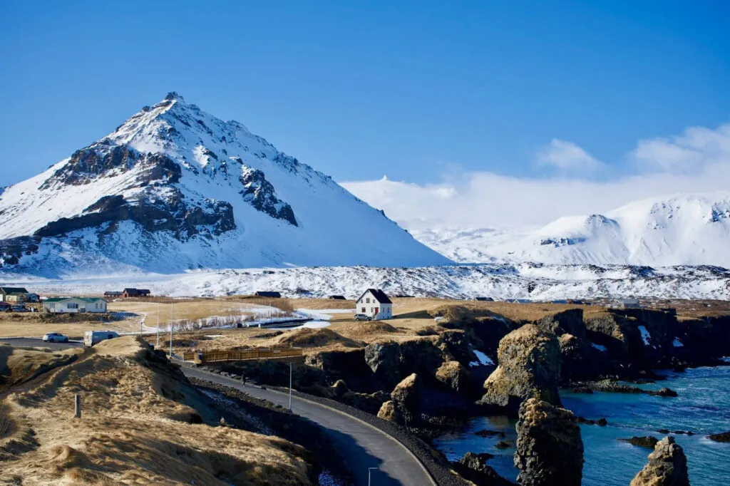 Landschapszicht op het schiereiland Snæfellsnes in IJsland, met een klein wit huisje aan de ruige kustlijn op de voorgrond en de majestueuze, met sneeuw en ijs bedekte vulkaangletsjer Snæfellsjökull onder een strakblauwe lucht op de achtergrond.