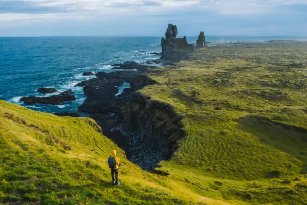 Een alleenstaande wandelaar in een opvallende gele jas staat op een groene klif en kijkt uit over de ruige basaltformaties van Lóndrangar en de blauwe oceaan aan de kust van Snæfellsnes, IJsland.