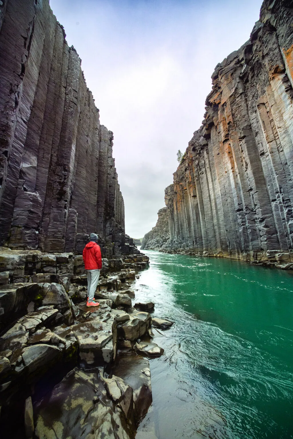 Een reiziger in een rode jas staat op de rotsen langs het groene water van de Stuðlagil Canyon in IJsland.