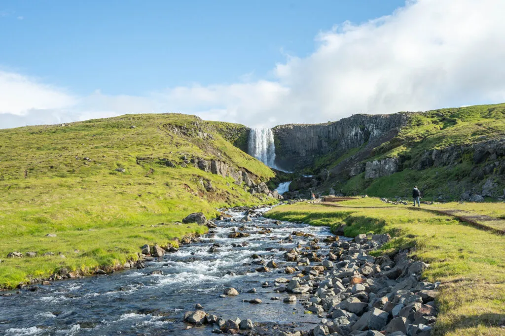 Een kabbelende rivier stroomt over stenen met op de achtergrond de Svartifoss waterval in het groene berglandschap van Vatnajökull Nationaal Park in IJsland.
