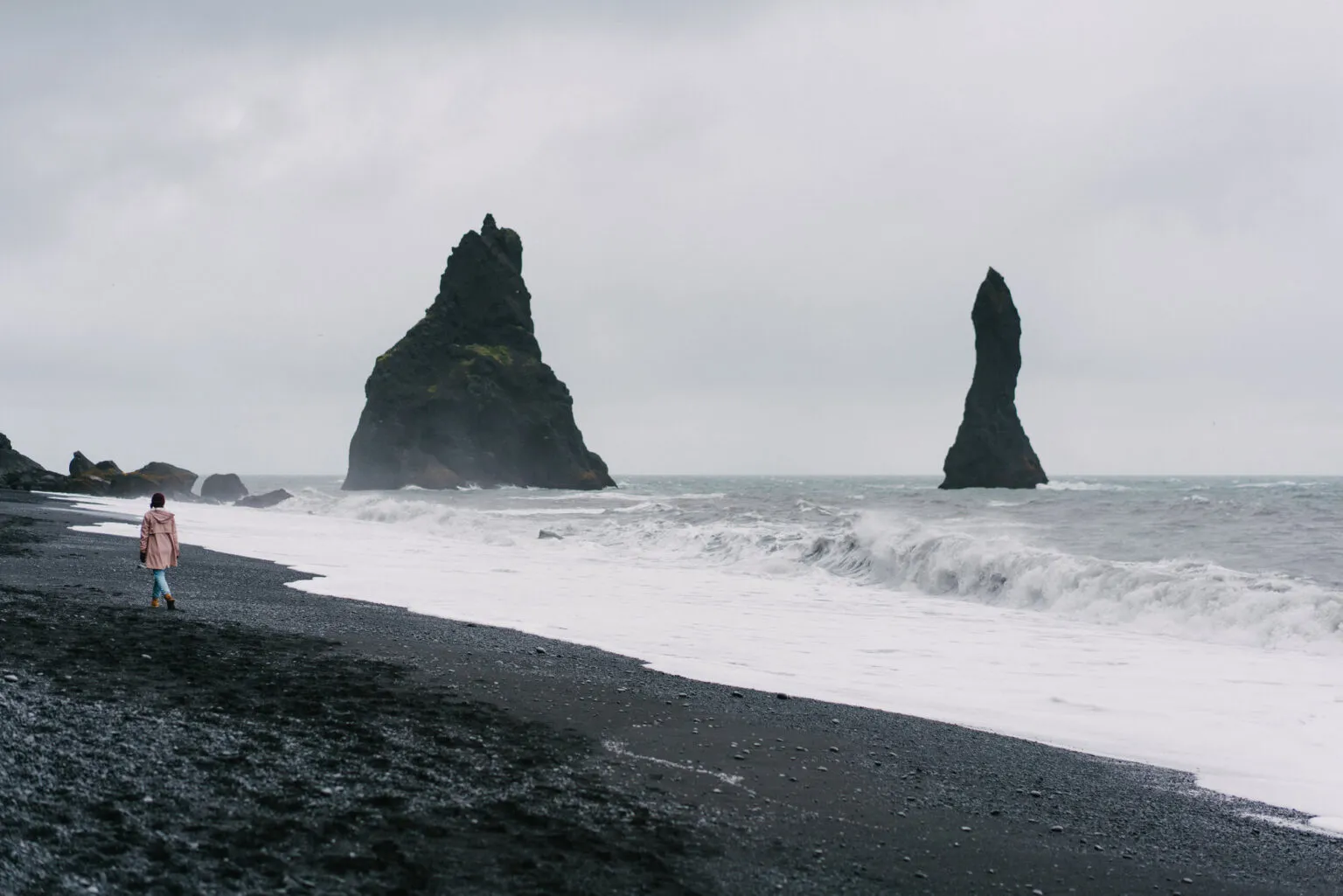 Een reizigster wandelt langs de schuimende branding op het zwarte vulkanische zandstrand van Reynisfjara bij Vík in IJsland, met de karakteristieke Reynisdrangar-rotsnaalden die uit de oceaan omhoogrijzen onder een mistige hemel.
