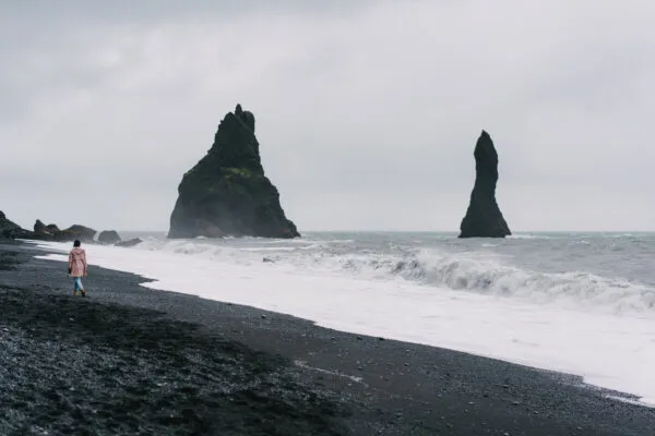 Een reizigster wandelt langs de schuimende branding op het zwarte vulkanische zandstrand van Reynisfjara bij Vík in IJsland, met de karakteristieke Reynisdrangar-rotsnaalden die uit de oceaan omhoogrijzen onder een mistige hemel.
