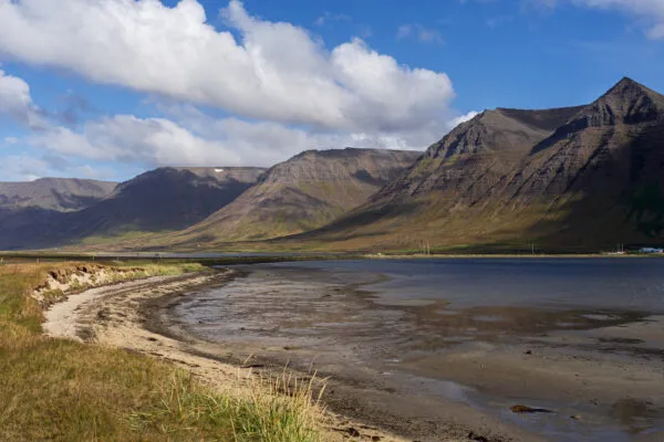 Zicht op het zandstrand van de Grundarfjörður-fjord in de IJslandse Westfjorden, met kleine rode huizen aan de voet van indrukwekkende, steile bergen en een boot die rustig op het water ligt.