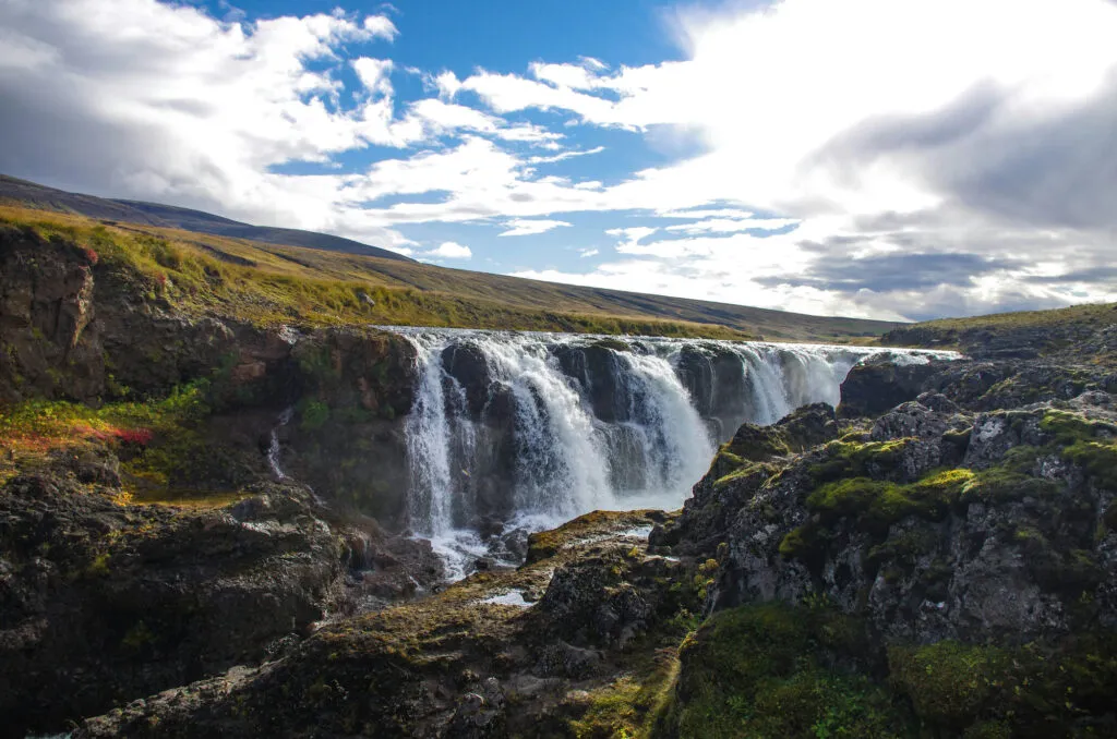 Zicht op de Kolufossar-waterval die door de diepe, rotsachtige Kolugljúfur-kloof stroomt in IJsland, onder een blauwe lucht met witte wolkpartijen.
