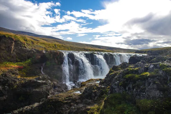 Zicht op de Kolufossar-waterval die door de diepe, rotsachtige Kolugljúfur-kloof stroomt in IJsland, onder een blauwe lucht met witte wolkpartijen.