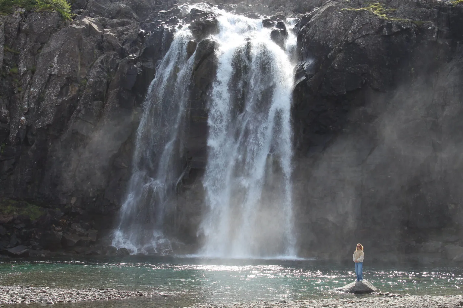 Een brede, krachtige waterval klettert langs een steile donkere rotswand naar beneden in een helder waterbekken, met een persoon die op de oever staat voor de opstijgende nevel in Fossfjörður, IJsland.