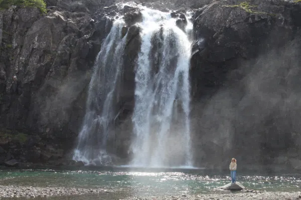 Een brede, krachtige waterval klettert langs een steile donkere rotswand naar beneden in een helder waterbekken, met een persoon die op de oever staat voor de opstijgende nevel in Fossfjörður, IJsland.
