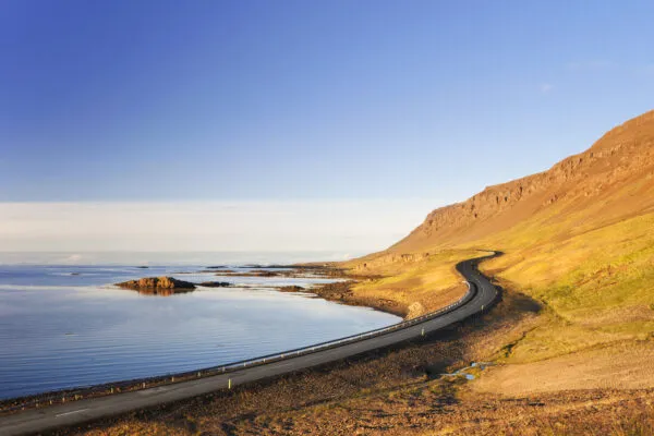 Een lege, geasfalteerde weg meandert langs de rustige blauwe kustlijn en de voet van een zonnige, steile berghelling in IJsland onder een helderblauwe hemel.