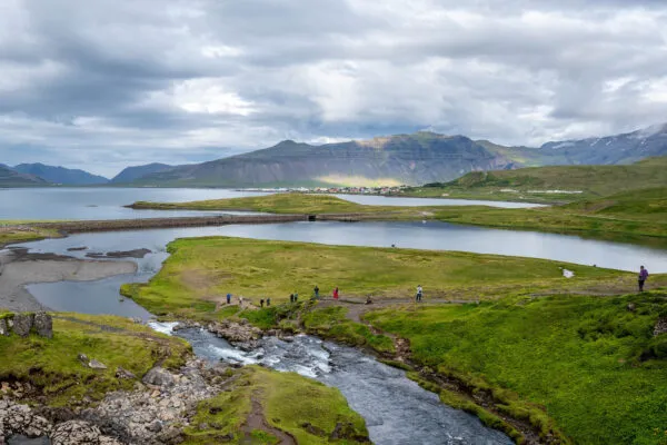 Uitzicht vanaf een verhoging op een stromende rivier die uitmondt in een baai, omgeven door groene weides met wandelende mensen en majestueuze bergen onder een bewolkte hemel in de Westfjorden van IJsland.