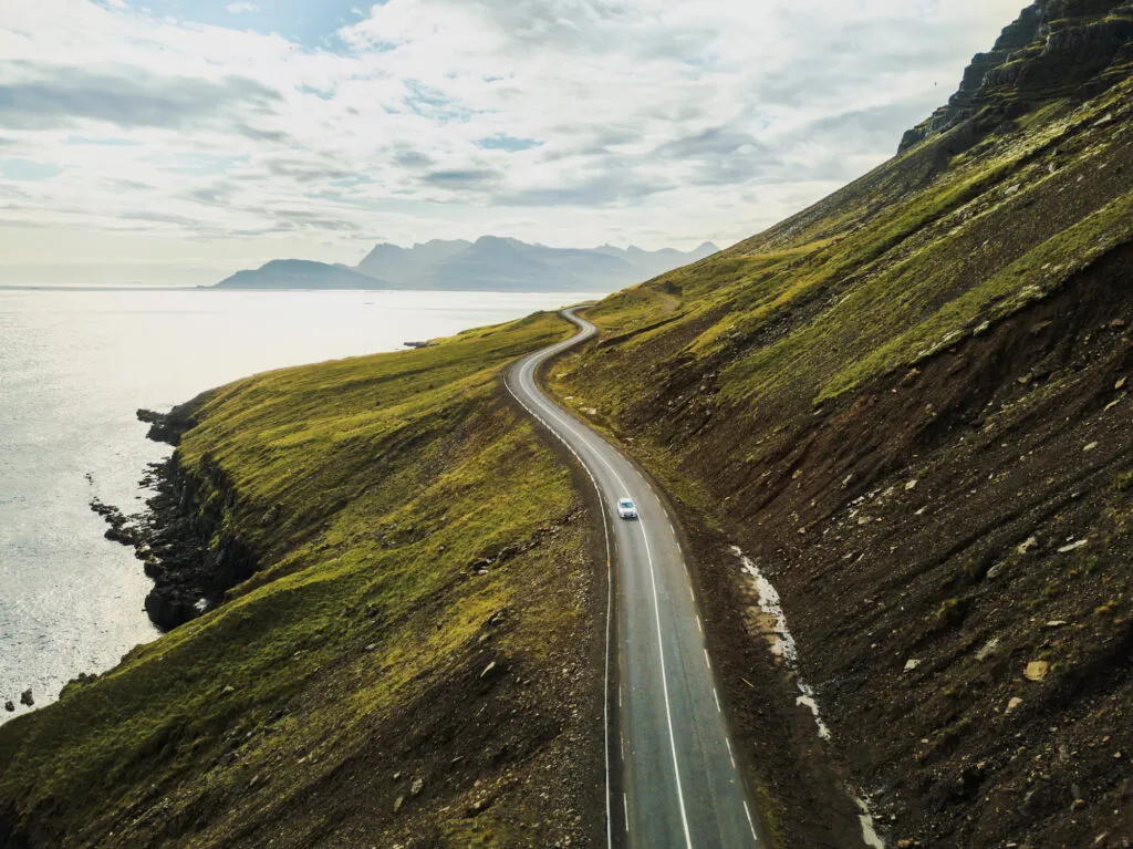 Een auto rijdt op een kronkelende weg langs groene bergen en de oceaan tijdens een roadtrip door IJsland.