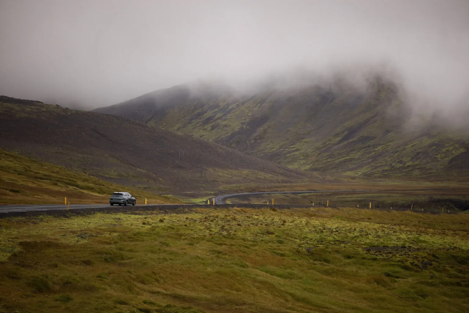 Een enkele auto rijdend over een asfaltweg door een uitgestrekt groen-bruin landschap met mistige, in wolken gehulde bergen op de achtergrond in IJsland.