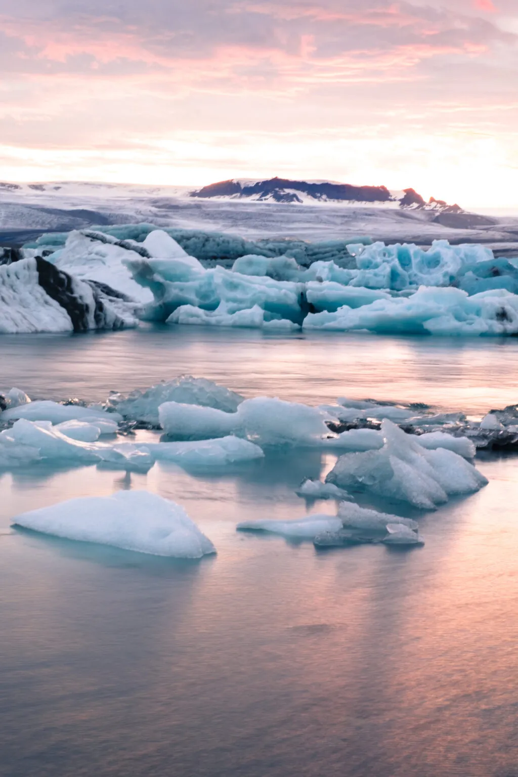 Blauwe en witte ijsbergen drijven in het kalme water van een gletsjerlagune onder een zachte avondlucht in IJsland.