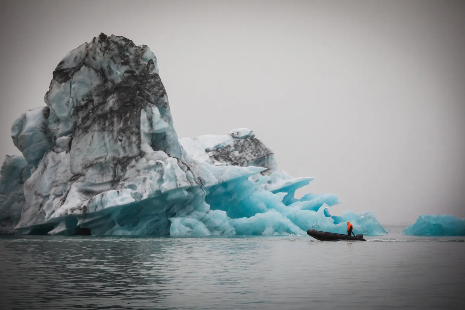 Een kleine zwarte RIB-boot met een passagier vaart op het kalme water vlak langs een enorm, helderblauw ijsbergmassief in de gletsjerlagune Jökulsárlón in IJsland.