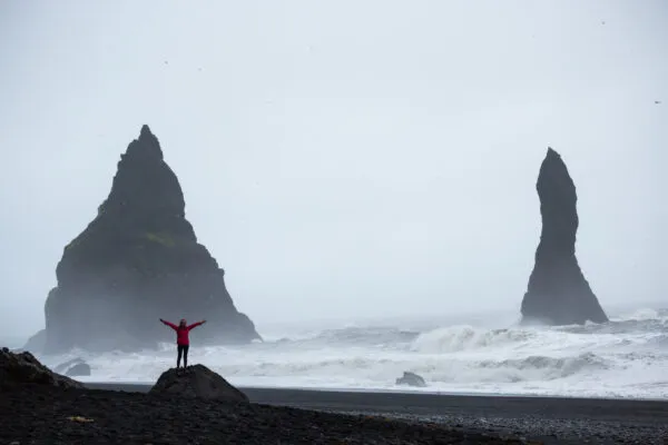 Een reiziger staat op een rots op het zwarte zandstrand van Reynisfjara met de armen wijd open naar de ruwe oceaan, met de imposante basaltrotsen van Reynisdrangar op de achtergrond.