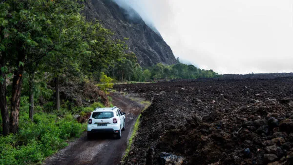 Een witte auto rijdt over een onverharde weg met aan de ene zijde groene bomen en aan de andere zijde een uitgestrekt landschap van donkere vulkanische lava op Fogo, Kaapverdië.