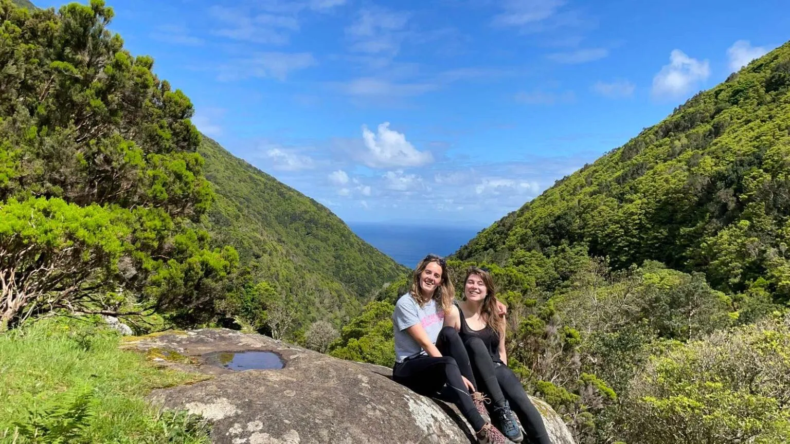 Twee vriendinnen zitten glimlachend op een grote rots in een zonovergoten groen berglandschap, met op de achtergrond een dal dat uitkijkt op de blauwe zee.