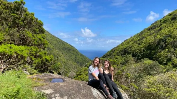 Twee vriendinnen zitten glimlachend op een grote rots in een zonovergoten groen berglandschap, met op de achtergrond een dal dat uitkijkt op de blauwe zee.