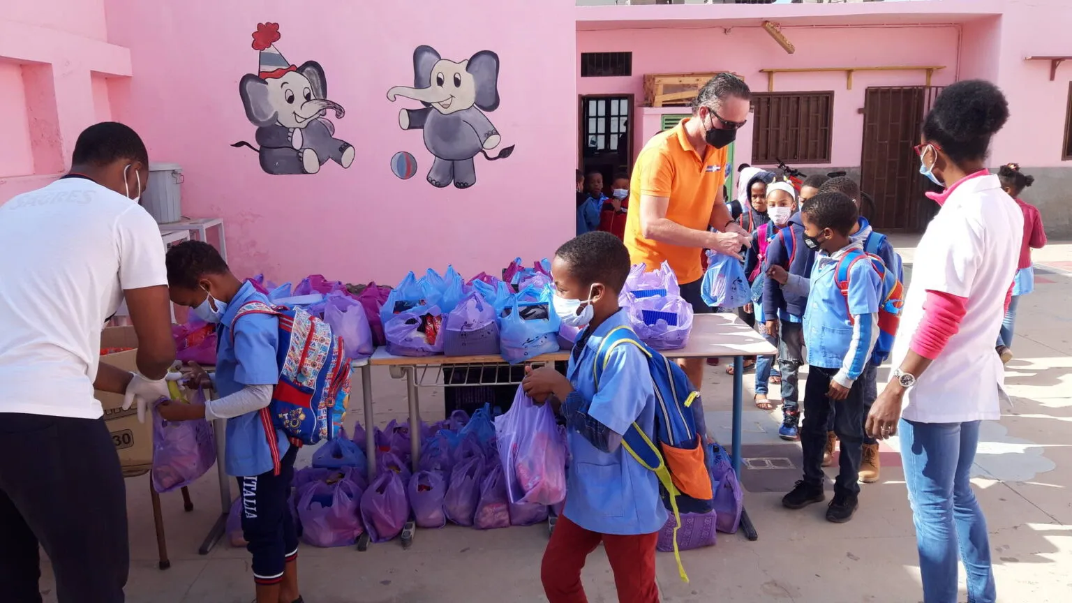 Vrijwilligers delen blauwe en paarse schooltassen uit aan basisschoolleerlingen in een klaslokaal met roze muren in Kaapverdië.