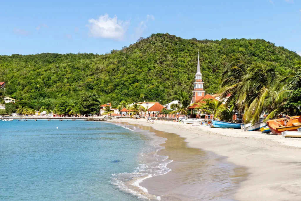 Zicht op het zandstrand en de rustige zee van Les Anses-d'Arlet op Martinique, met op de achtergrond de opvallende kerktoren, kleurrijke huisjes en groenbegroeide bergen onder een heldere blauwe lucht.