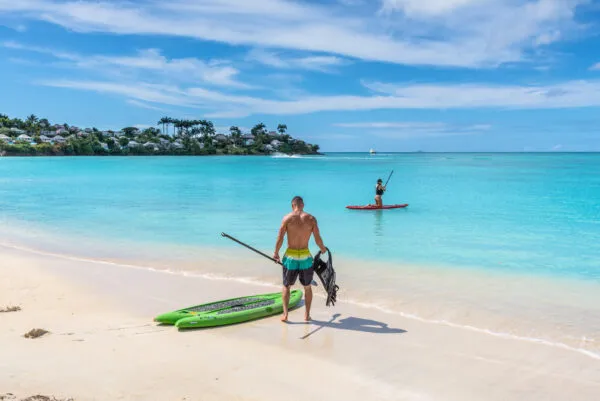 Een man staat met zijn supplank op het witte zandstrand van Valley Church Beach op Antigua, terwijl een vrouw verderop op het turquoise water aan het suppen is onder een heldere hemel.