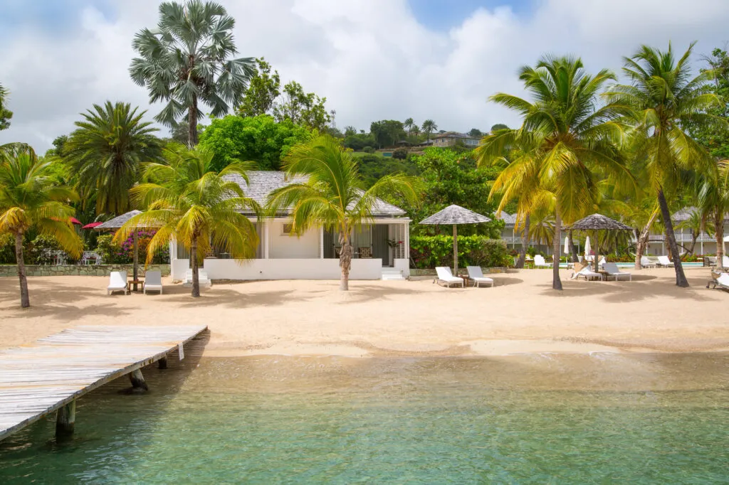 Zicht op een idyllisch zandstrand van een resort op Antigua met palmbomen, ligbedden en parasols, grenzend aan rustig, helder water en een klein stukje van een houten steiger op de voorgrond.