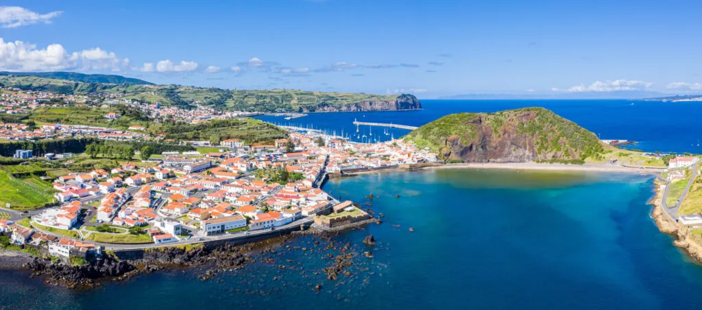 Hoog zicht op de stad Horta met rode daken, een zandstrand en een beschutte, helderblauwe baai tegen groene heuvels.