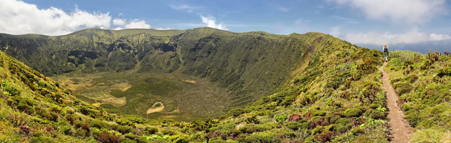 Panorama van een zandpad op de rand van een immense, diepe groene vulkaankrater onder een licht bewolkte lucht.