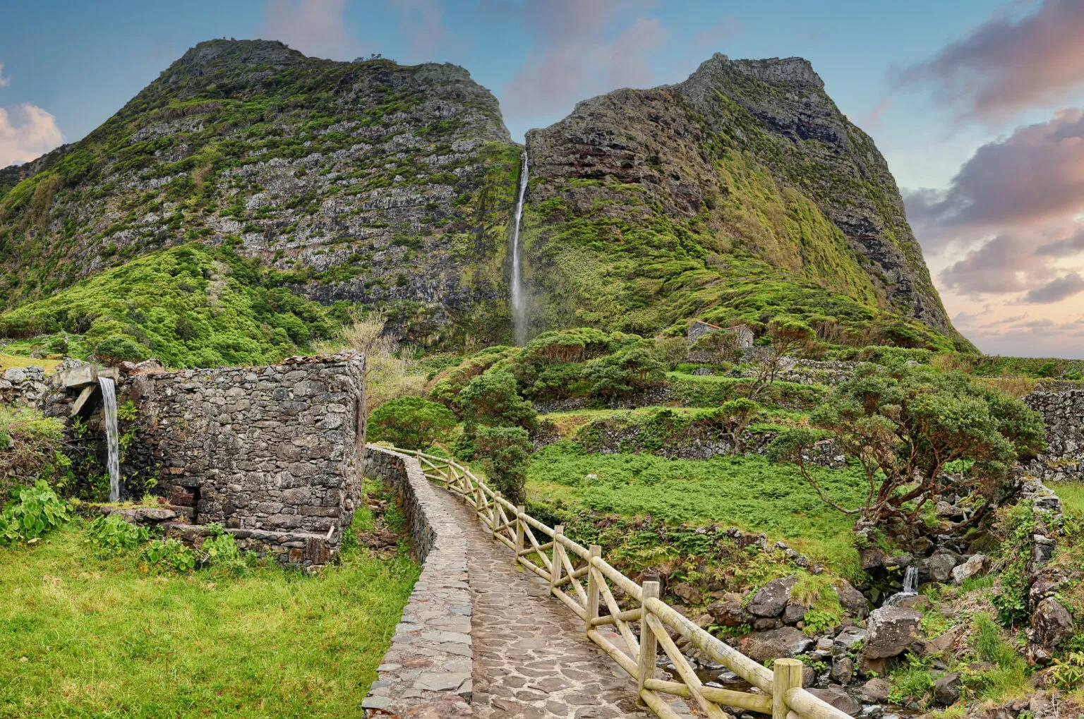 Een stenen wandelpad met houten hekwerk leidt naar een hoge waterval die langs een steile, groene bergwand naar beneden valt.