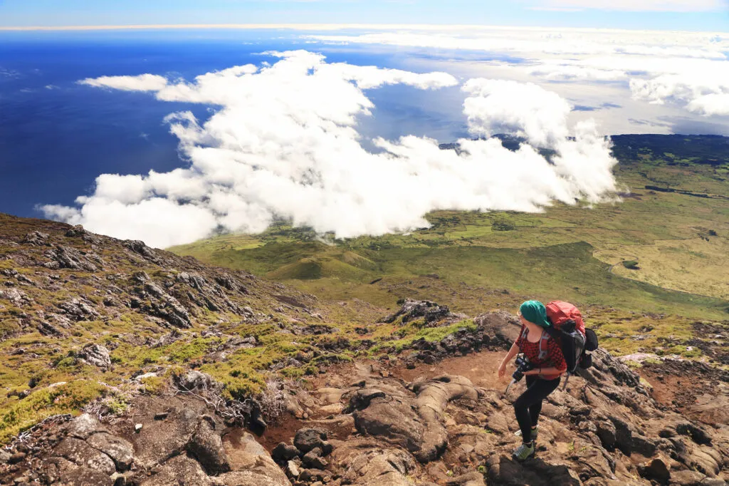 Een hiker met een grote rugzak klimt over het steile lavagesteente van Mount Pico met uitzicht op het wolkendek en de oceaan.