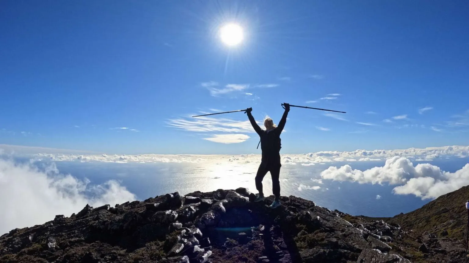 Silhouet van een wandelaar die zegevierend met wandelstokken in de lucht op de bergtop van Mount Pico staat, met een zon overgoten uitzicht boven de wolken op de Azoren.