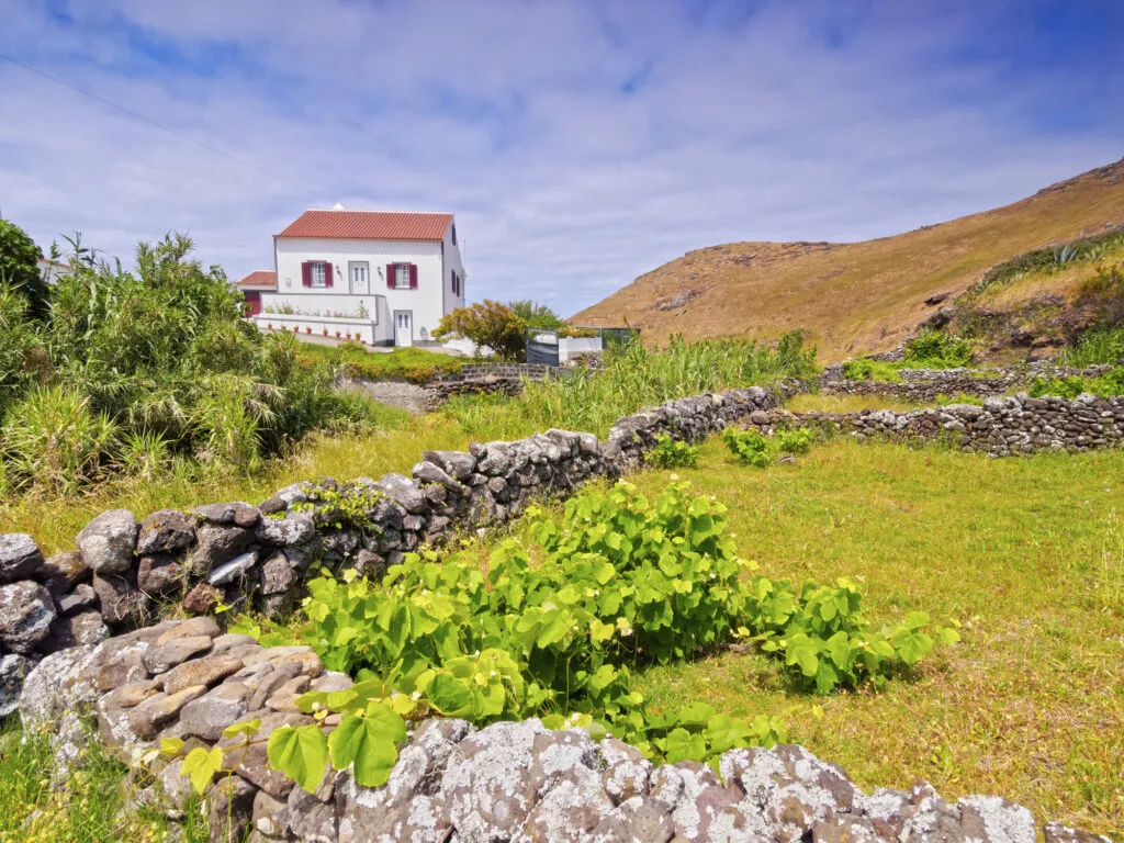 Een wit traditioneel huisje met een rood dak in een groen landschap met opgebouwde stenen muurtjes onder een blauwe lucht.