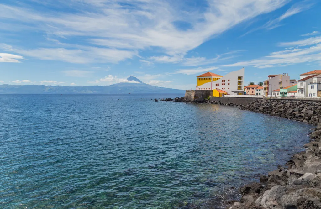 Zicht op de rotsachtige kust en witte gebouwen van het stadje Velas op São Jorge, met over het blauwe water de vulkaan Mount Pico aan de horizon.