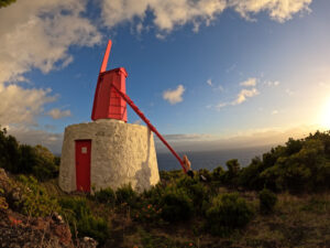 Een karakteristieke witte stenen windmolen met een felrode houten kap en wiekenas aan de kust van São Jorge bij zonsondergang.