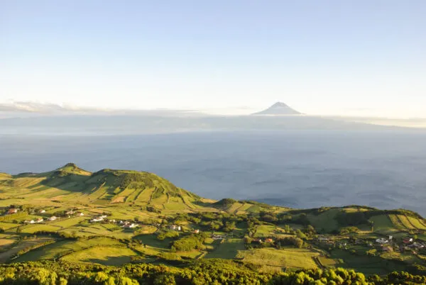 Een weids uitzicht over de groene, glooiende hellingen van São Jorge met de blauwe oceaan en de markante piek van de vulkaan Mount Pico op de achtergrond.